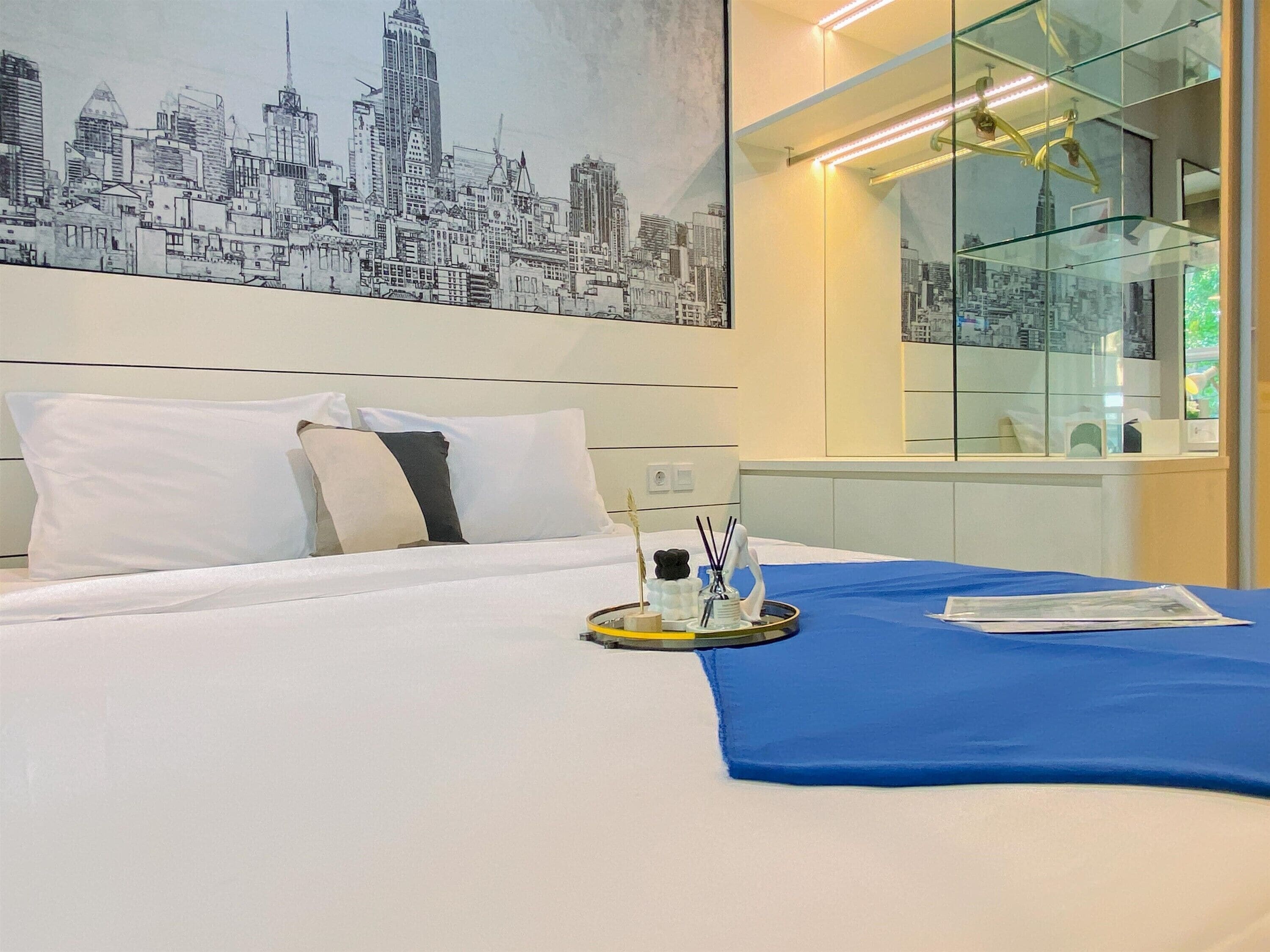 Une chambre d’hôtel avec un lit, un tableau représentant un paysage urbain accroché au mur, une cloison en verre et un élément décoratif bleu sur le lit.