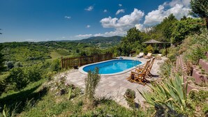A swimming pool with a wooden deck, surrounded by lush greenery and a scenic view of hills.