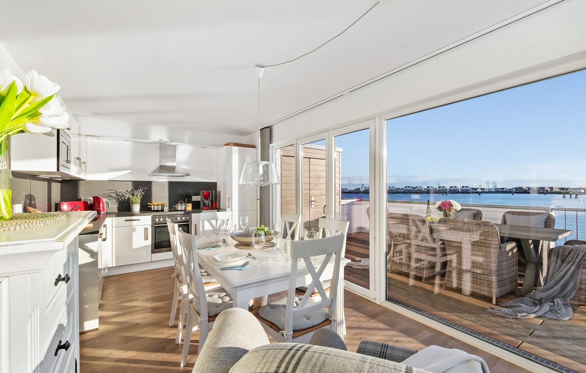 Une cuisine moderne avec des meubles blancs et un coin repas comprenant une table et des chaises. De grandes fenêtres offrent une vue sur la mer.