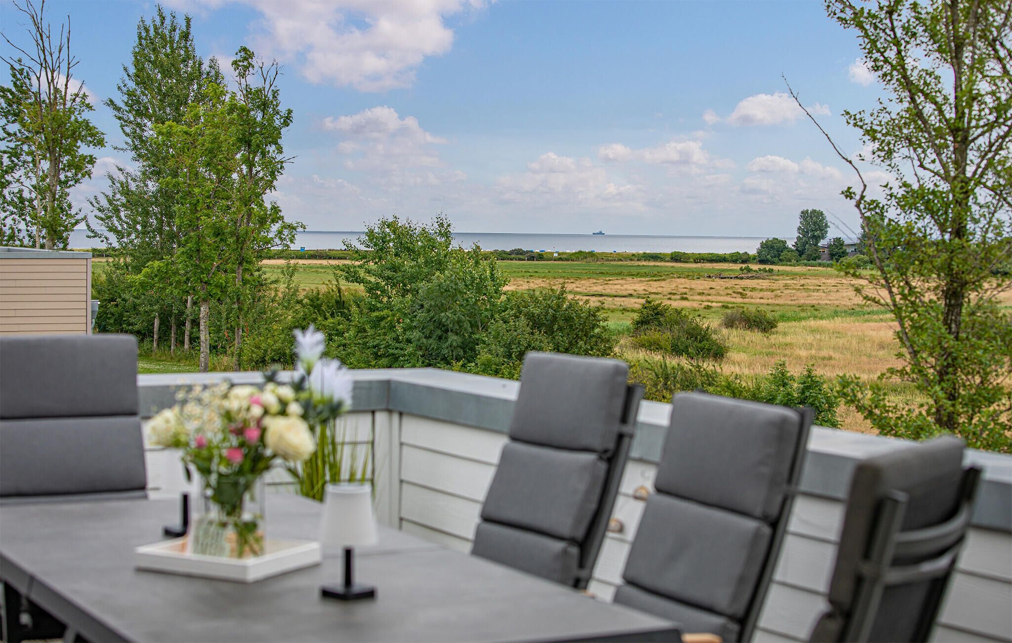 Eine Terrasse mit Tisch und Stühlen, einem Blumenstrauß und Blick auf ein Feld und ein Gewässer.