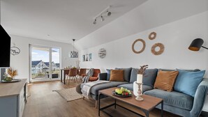 Een moderne woonkamer met een grijze bank, een salontafel met fruit, een eethoek met stoelen en een groot raam met uitzicht op huizen.