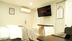 Moderni hotellihuone, jossa on sänky, työpöytä kannettavalla tietokoneella, seinälle asennettu televisio ja seinään kiinnitetty ilmastointilaite.