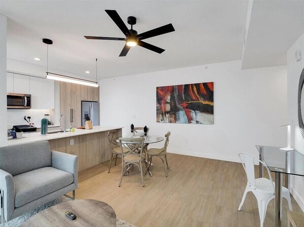 A modern kitchen with white cabinets, a microwave, and a coffee machine. There is a dining area with a glass table and chairs, and a gray sofa. A ceiling fan is visible above the dining area.
