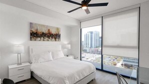 A modern hotel room with a large bed, white bedding, a painting on the wall, a ceiling fan, and a view of the city through large windows. - Central Studio | Pool/Spa/Hottub | Close to Shops & Parks (Phoenix)