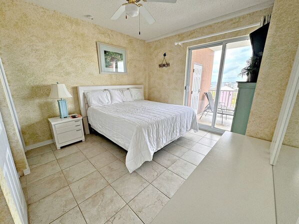 A bedroom with a bed, a nightstand, a ceiling fan, a framed picture, and a view of the outside.