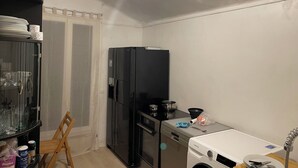 Una cucina compatta con frigorifero nero, lavastoviglie e lavatrice. Si notano una sedia, un tavolo con stoviglie e un sacchetto di plastica.