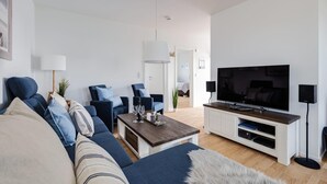 Moderni olohuone, jossa on sohva, nojatuoleja, sohvapöytä, televisio ja valkoinen mediakeskus.