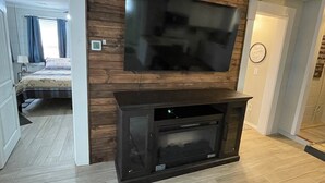 Een woonkamer met een flatscreen televisie aan een houten wand, een open haard met een televisie eronder, en een bed met een beddengoed met een patroon op de achtergrond.
