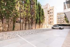 A paved courtyard with a car parked on the right, surrounded by buildings with balconies and a stone wall.