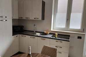 A compact kitchen with a microwave, sink, and wooden bowl on the counter, a dining table with a chair, and a window with blinds. - Cozy 36 sqm apartment in Trani, ideal for 2 people. Located on the ground floor, it includes a reserved outdoor parking space. Just a short walk from the center of Trani, it offers easy access to all the main attractions. Trani is a charming city in Pugl (Trani)