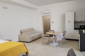 A modern, well-lit room with a sofa, dining table, chairs, and a kitchen area.