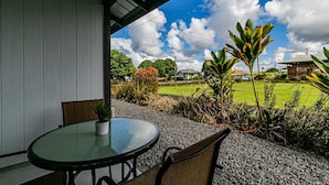 Een overdekt terras met een ronde tafel en stoelen, omringd door grind en planten, met uitzicht op een groen gazon en gebouwen.