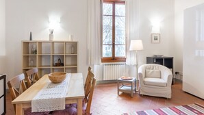 A room with a wooden dining table, chairs, a small side table, a white armchair, a bookshelf, a window with curtains, and a radiator. - GuestHost - Cozy and quiet apartment of 60 sqm, for 4 people, located on the 1st floor of a building (no elevator). The property is located near Piazza Dalmazia, from where the tram leaves to reach in a few minutes the Santa Maria Novella Station and the (Florence)