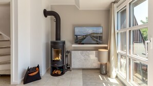 A modern living room with a wood-burning stove, a television mounted on a wall, and a large window providing natural light.