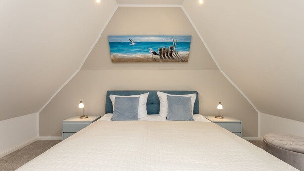A bedroom with a large bed, two bedside lamps, a painting of seagulls, and a view of the sky.