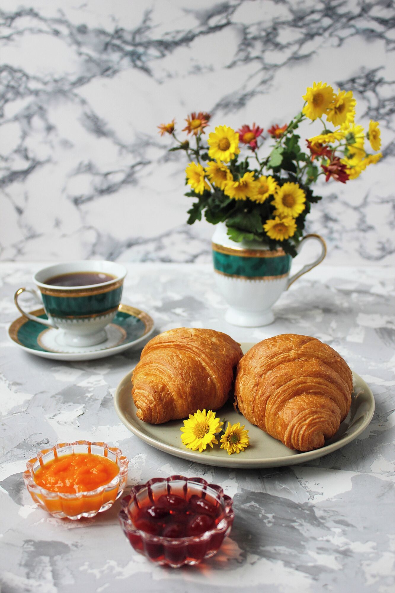 Une table en marbre sur laquelle se trouvent une assiette de croissants, une tasse de café et un vase rempli de fleurs jaunes.