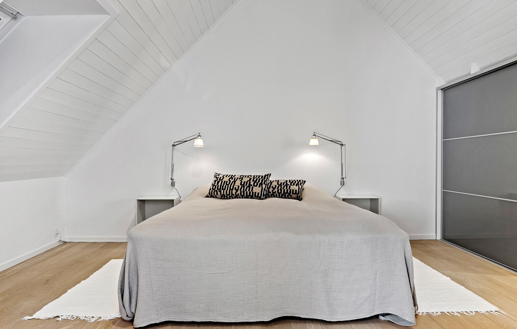 Ein Schlafzimmer mit schrägem Dach, einem Bett mit grauer Bettdecke, zwei Nachttischlampen und einem großen Fenster mit Jalousien.