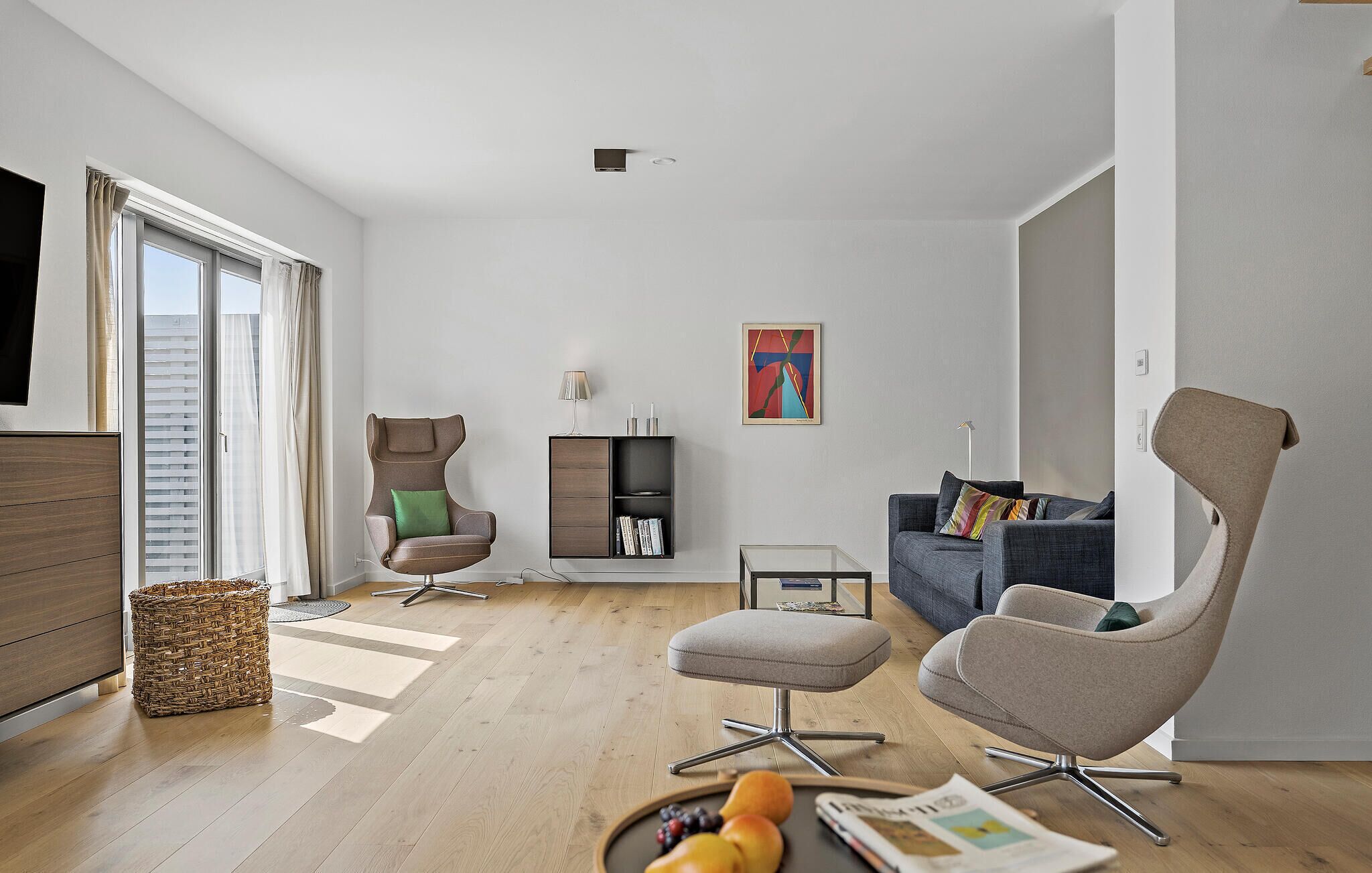 Una sala de estar moderna con piso de madera, un sofá gris, un sillón marrón, un pequeño reposapiés, una estantería y un televisor de pantalla plana.