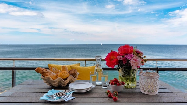 Mesa posta com pão, pratos, talheres, um vaso de flores, um copo de água, uma garrafa e uma cesta de morangos em um deck de madeira com vista para o mar e barcos.
