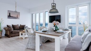 Ampla sala de estar com mesa de jantar para quatro pessoas, grandes janelas com vista para o mar, um sofá confortável e uma televisão montada na parede.