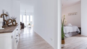 Quarto moderno e minimalista com piso de madeira, uma cama com colcha branca, uma mesinha pequena e uma janela grande.