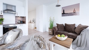 Uma sala de estar moderna com um sofá marrom, cadeiras de vime, uma mesa de centro de madeira com frutas e uma cozinha ao fundo.