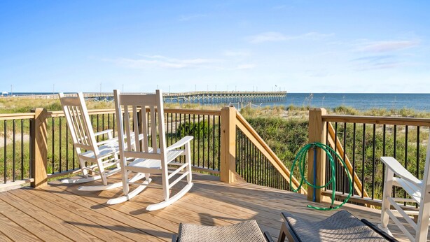 A wooden deck with white rocking chairs and a wicker lounge chair overlooking a beach with a pier in the distance.