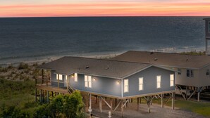 A beachfront house with a deck and multiple windows, elevated on stilts, overlooking the ocean.