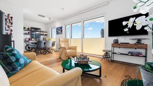 Un salon moderne avec un canapé, une table basse et un jeu d’échecs. On y trouve également un coin repas avec des chaises et une table, ainsi qu’une cuisine à l’arrière-plan.