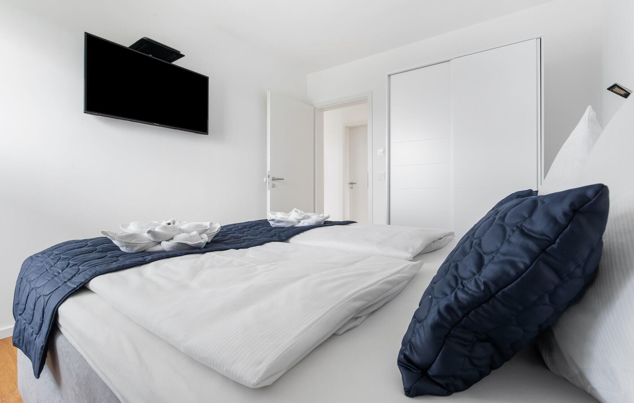 Quarto moderno com cama, paredes brancas, televisão fixada na parede e uma porta que dá para outro espaço.