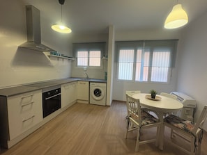Eine kompakte Küche mit Herd, Backofen, Spüle und Waschmaschine. Ein Esstisch mit Stühlen und ein Fenster mit Vorhängen sind ebenfalls vorhanden.