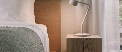 A bedroom with a wooden headboard, a bed with white bedding, a side table with a lamp, and sheer curtains.