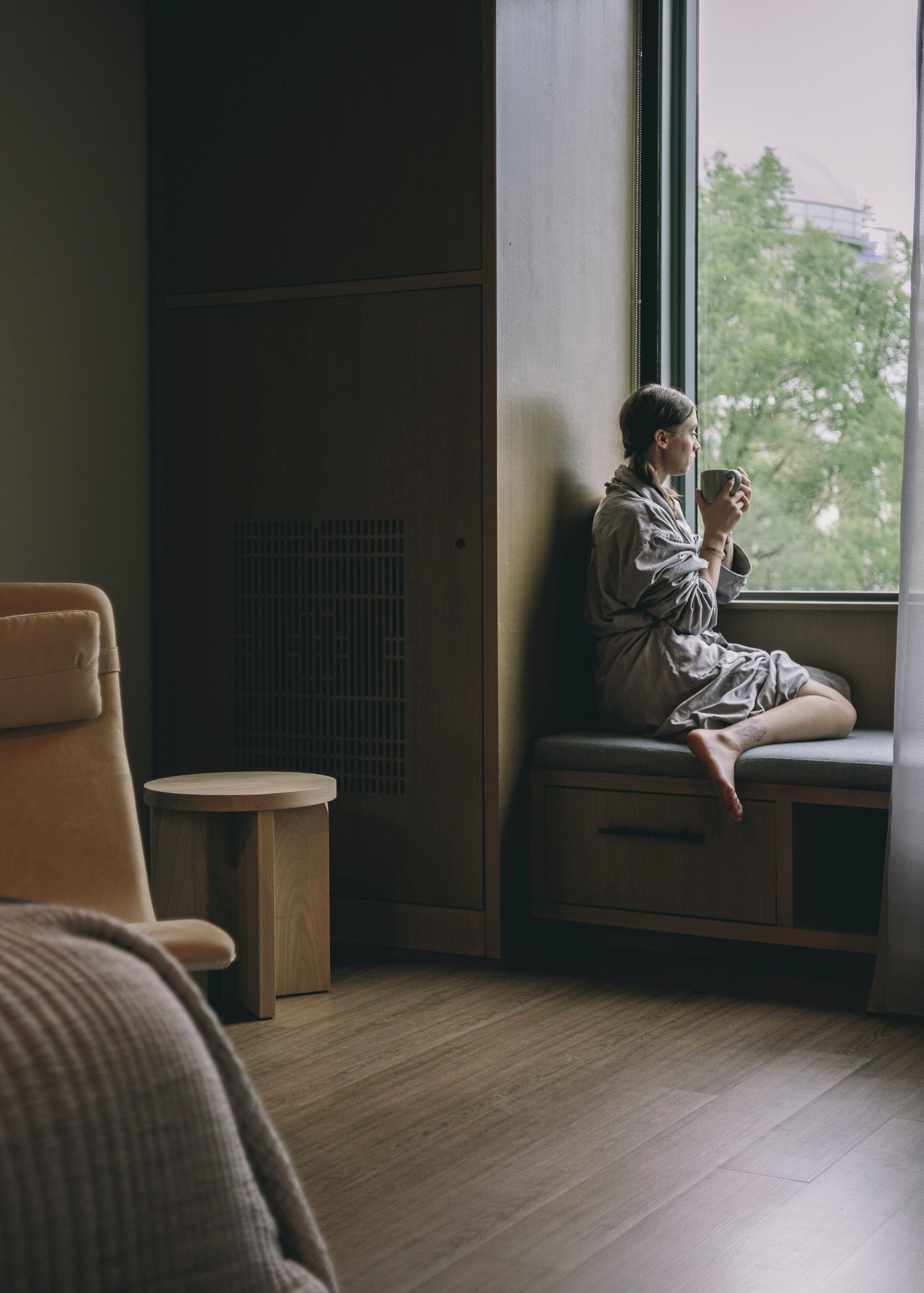 A person sitting by a window in a room with a wooden floor, a small round table, and a bench.