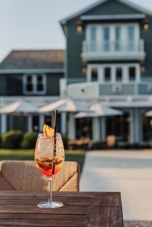 A cocktail glass with a lemon slice on a wooden table, with a large house in the background. - Stonebreaker Hotel (Fayetteville)