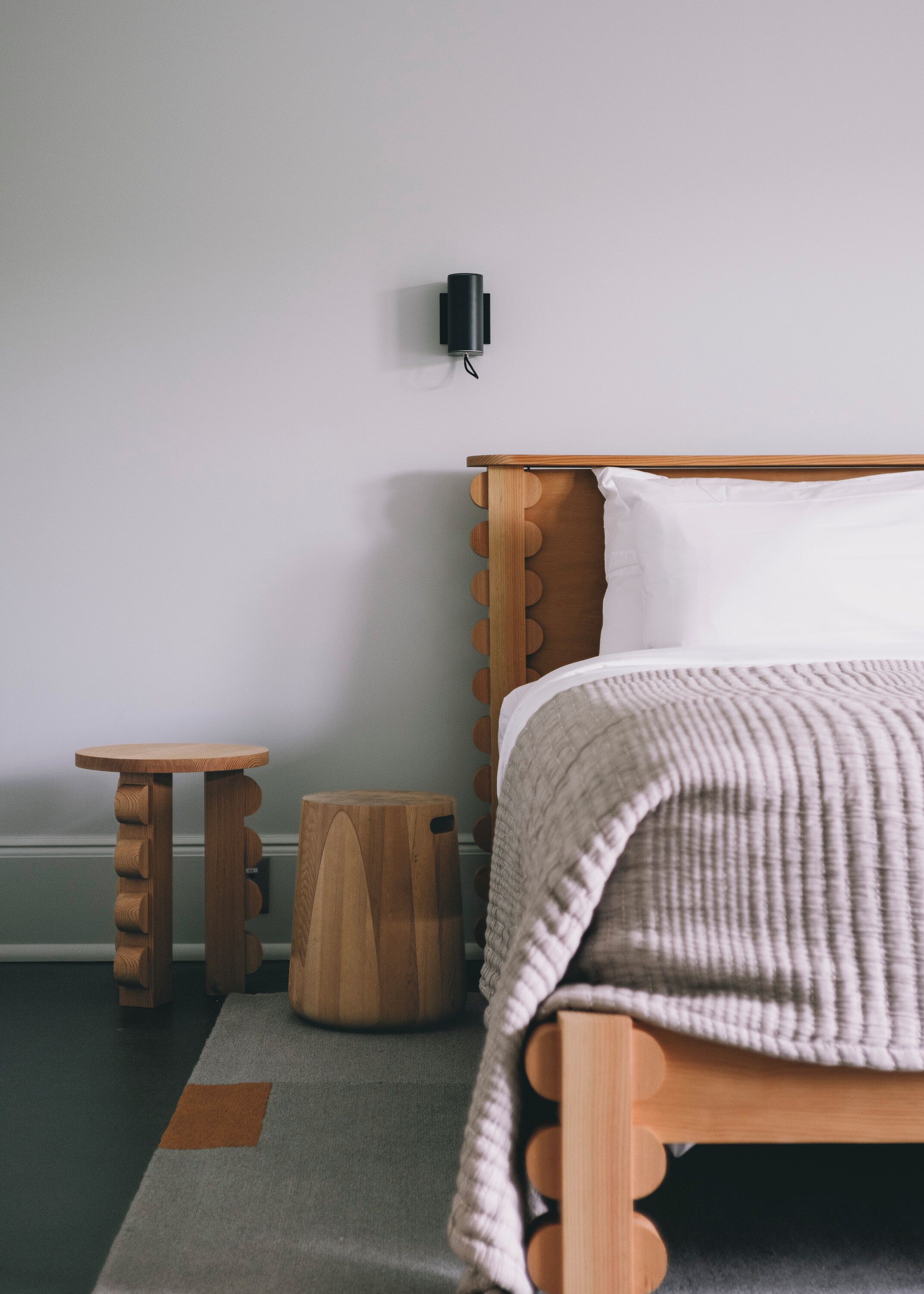 A bedroom with a wooden bed, a small wooden nightstand, a round wooden stool, and a wall-mounted light fixture.