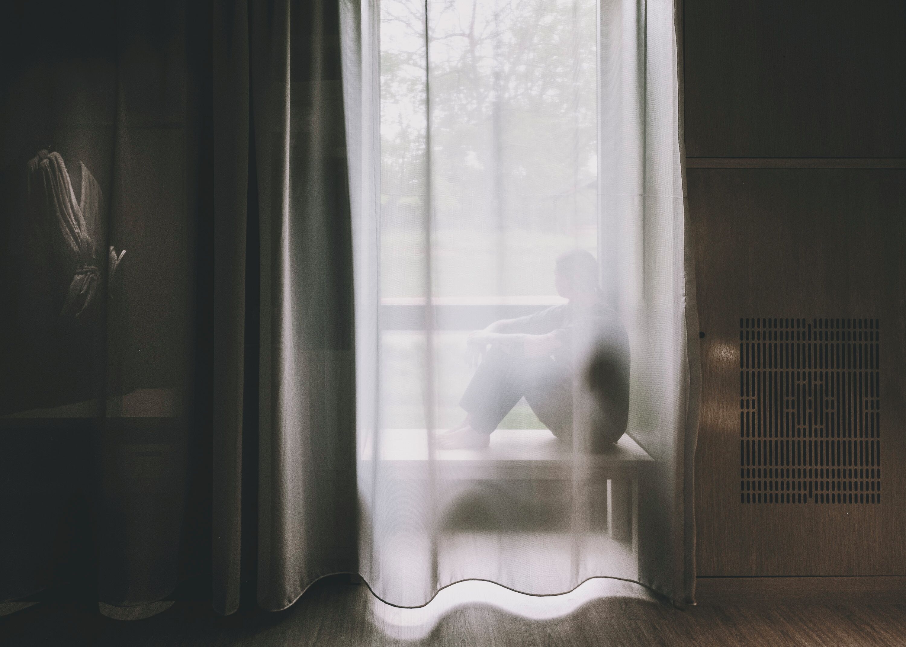 A room with a large window covered by sheer curtains, a radiator, and a wooden floor.
