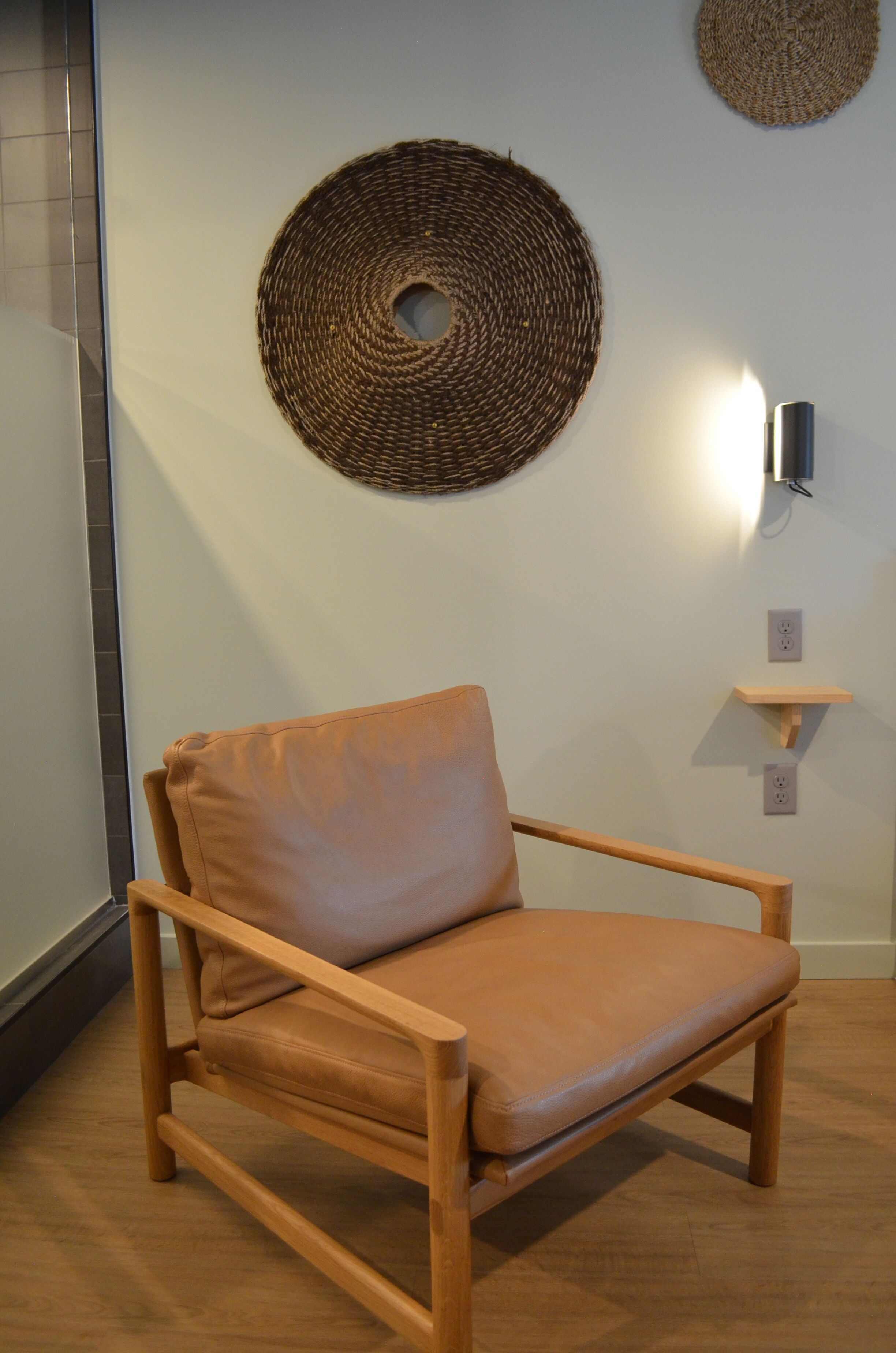 A wooden chair with a leather cushion in a room with a woven wall hanging and a small shelf.