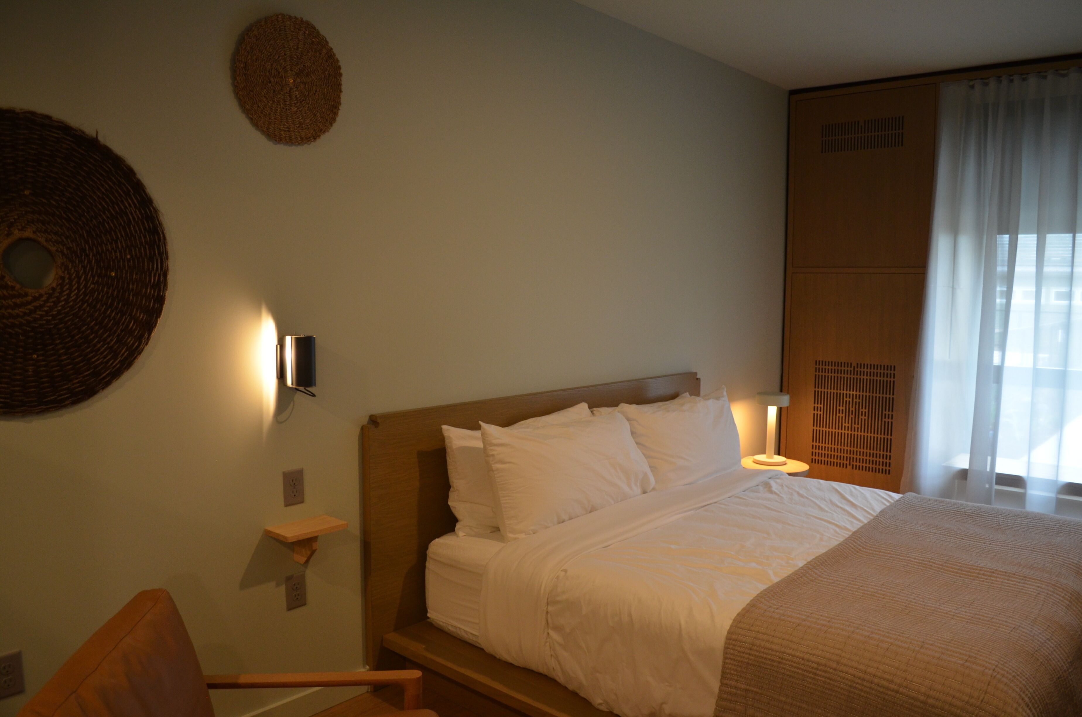 Una cama bien hecha con ropa blanca, una lámpara de noche, un cabecero de madera y un adorno de pared tejido.