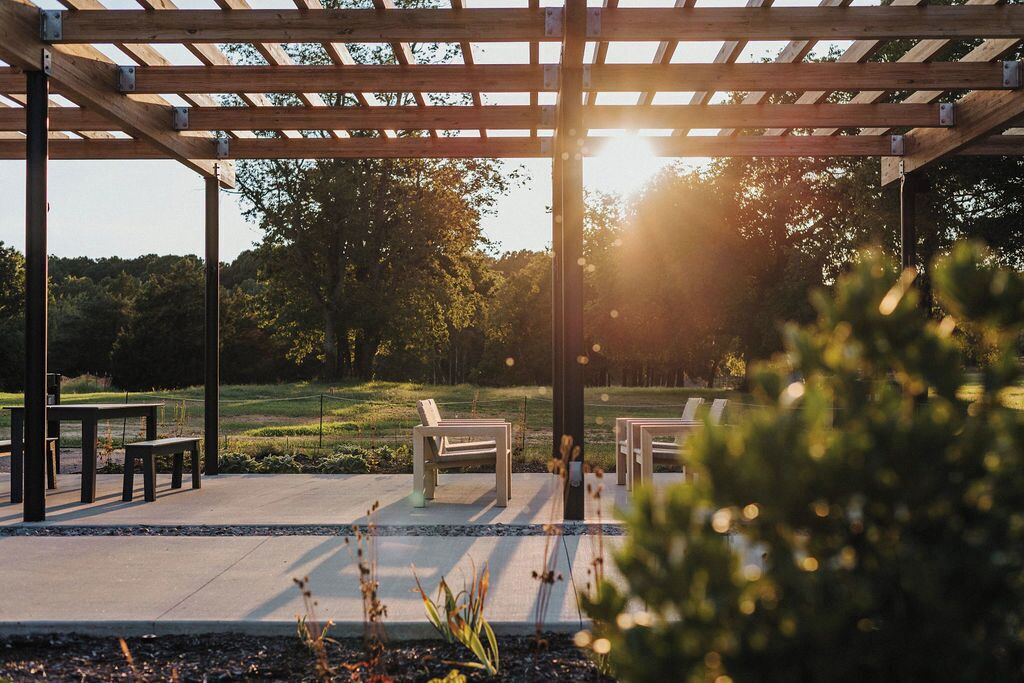 A wooden pergola structure with benches and tables, set in a garden with trees and a clear sky.