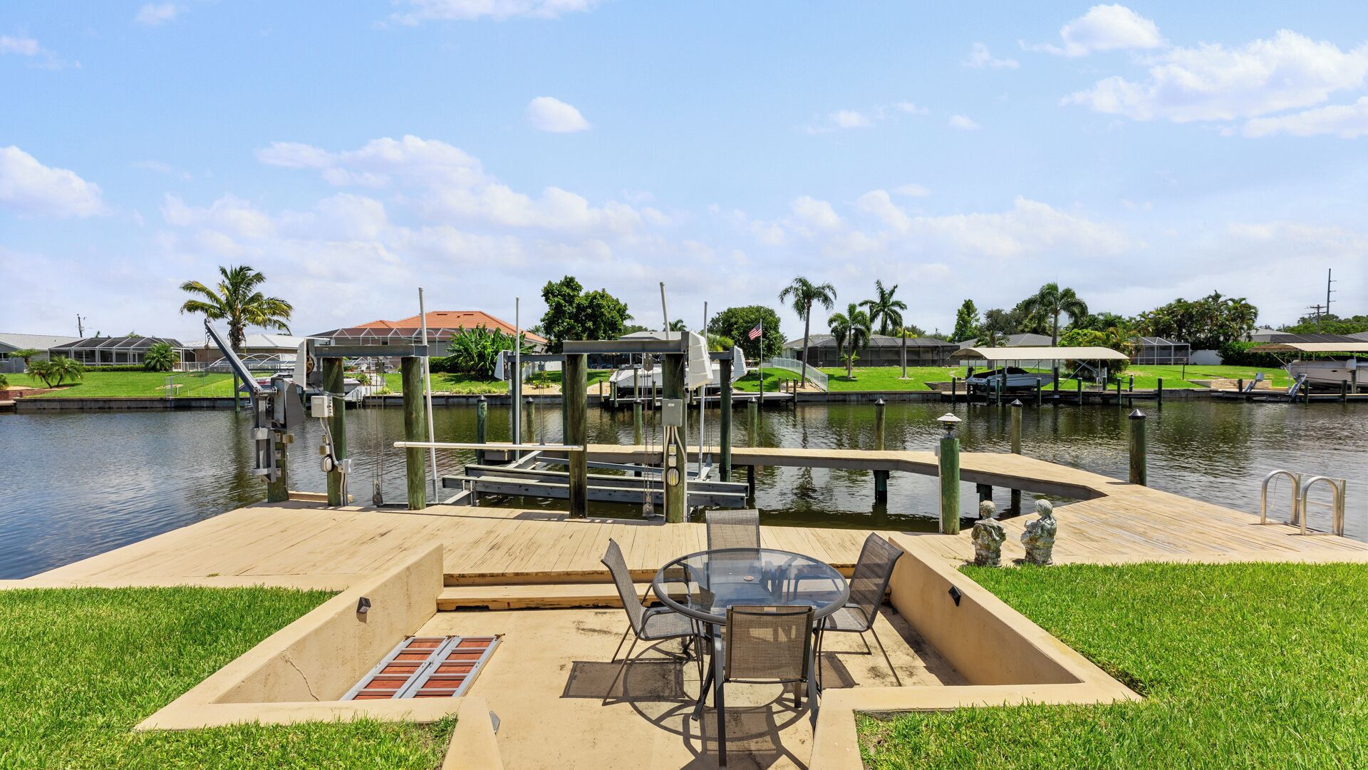 Une terrasse au bord de l’eau, avec une table et des chaises, un quai avec des bateaux, et une vue sur l’eau et la végétation environnante.