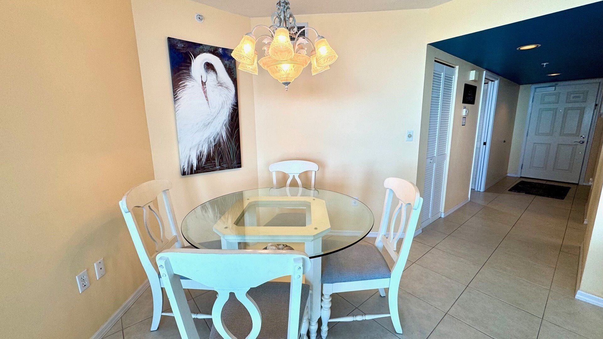 Un comedor con una mesa de vidrio y sillas blancas, una araña y un cuadro de un cisne en la pared.