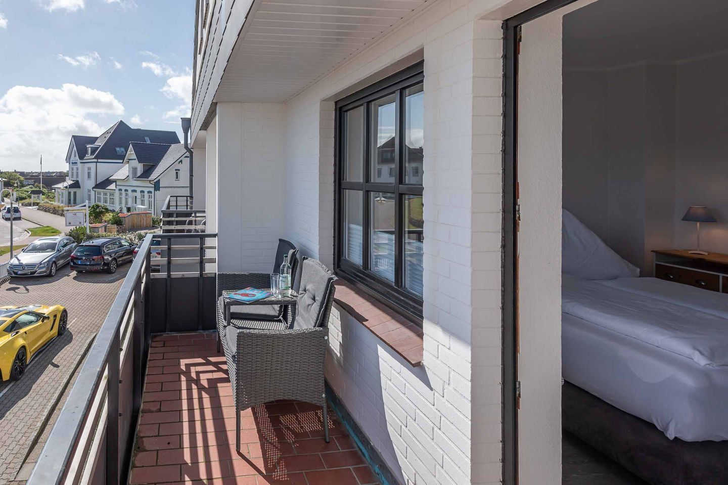 A balcony with wicker furniture, a bed, and a view of parked cars and houses.