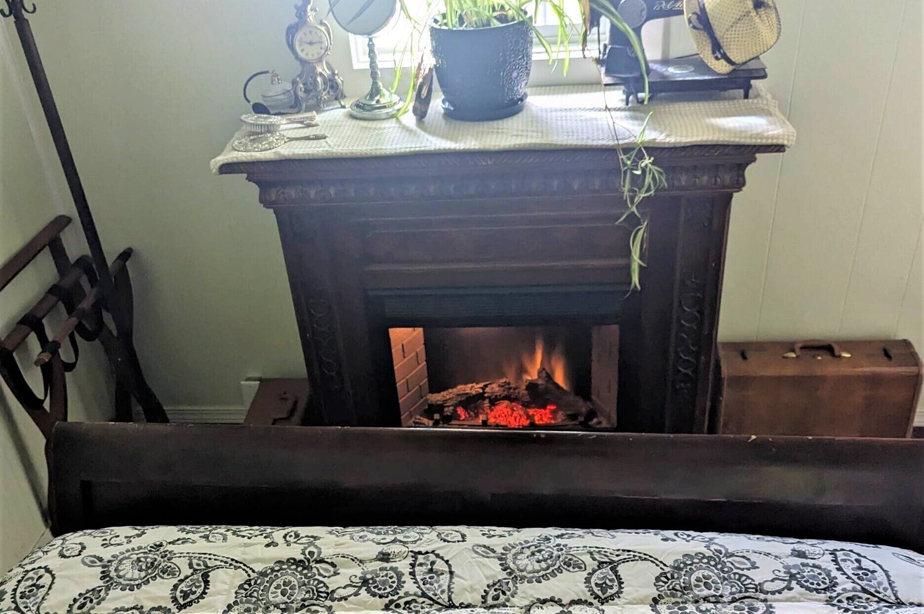 A room with a fireplace, a bed with a patterned bedspread, a chair, and a table with various items including a clock and a plant.