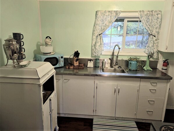Una cocina con armarios blancos, un lavamanos, un microondas, una tetera y un ventanal con cortinas estampadas.
