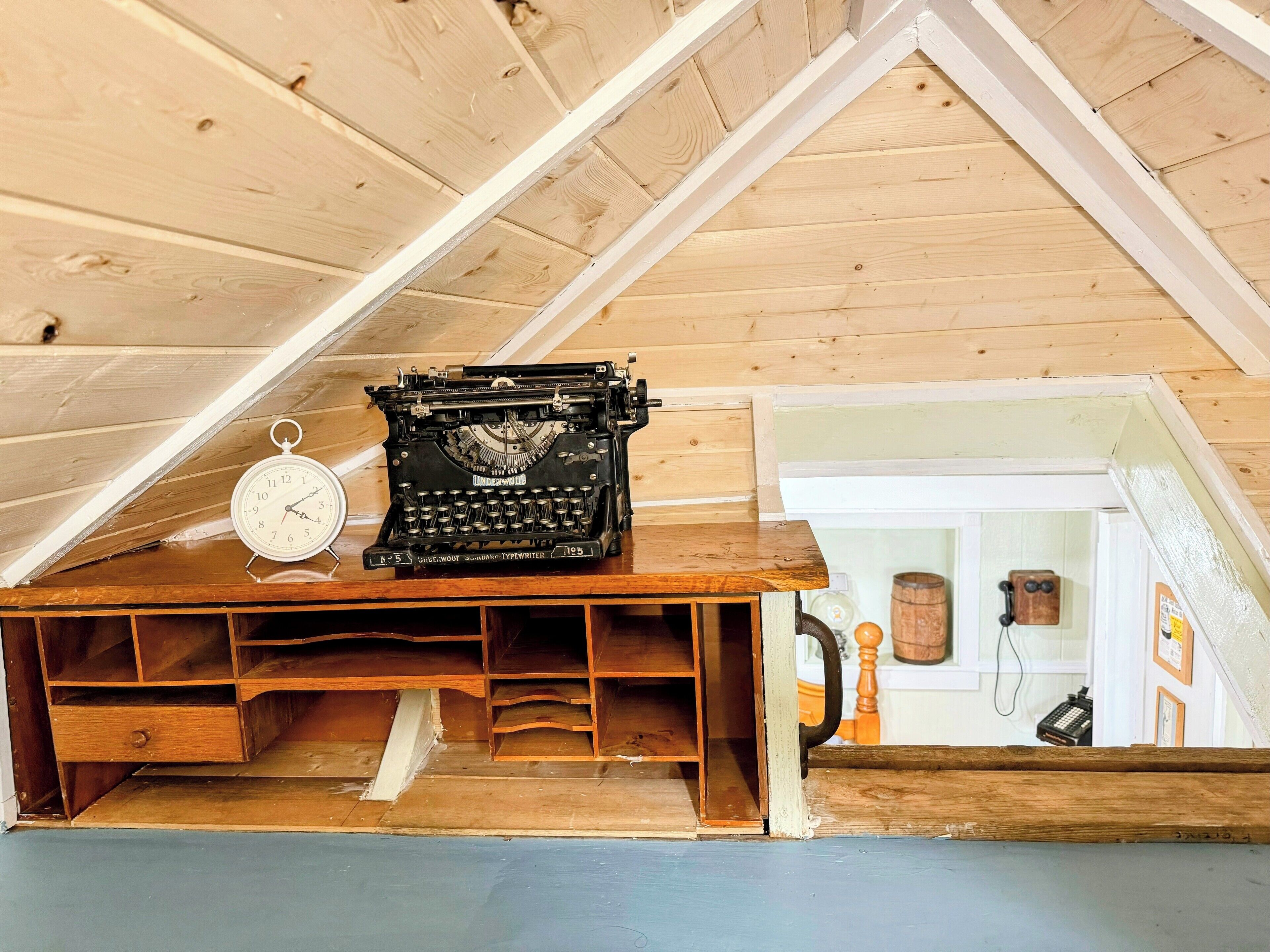 A wooden desk with a typewriter and a clock in a room with wooden walls and a sloped ceiling.
