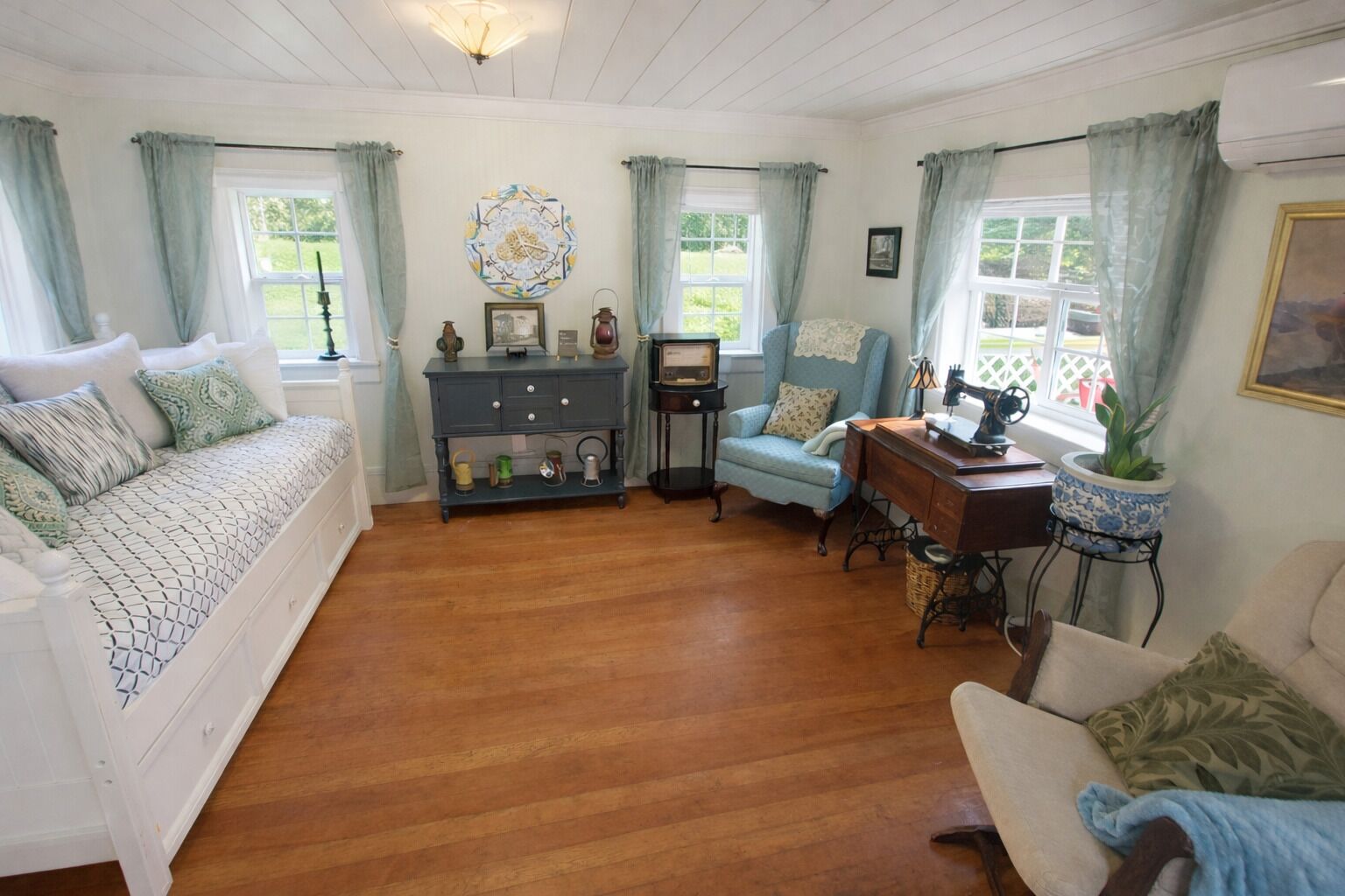 Een gezellige kamer met een houten vloer, een bank, een stoel, een bureau met een naaimachine en een dressoir.
