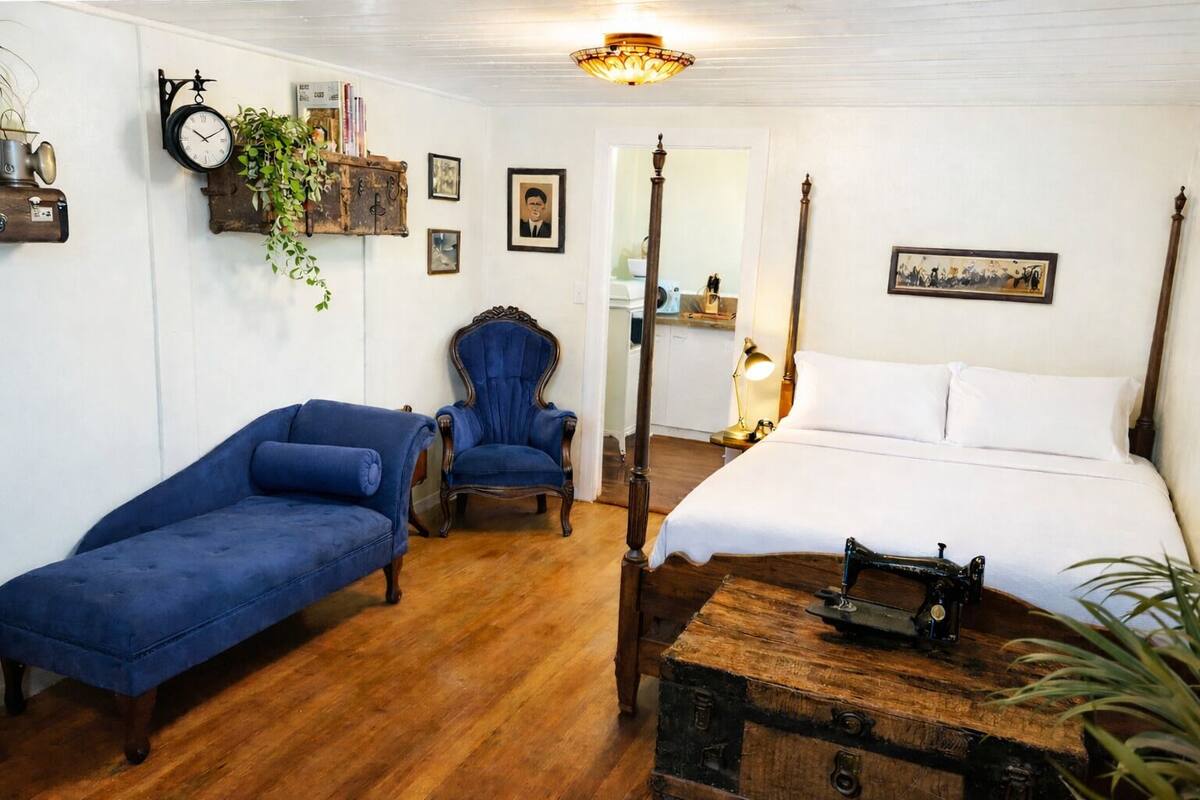Ein Schlafzimmer mit einem Himmelbett, einem blauen Sessel, einer Holzkommode und einer Vintage-Uhr an der Wand.