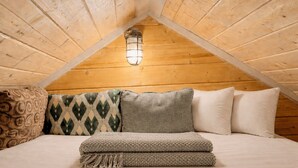 Een gezellig zolderkamer met een schuin houten plafond, een enkel bed met veel kussens en een hanglamp.