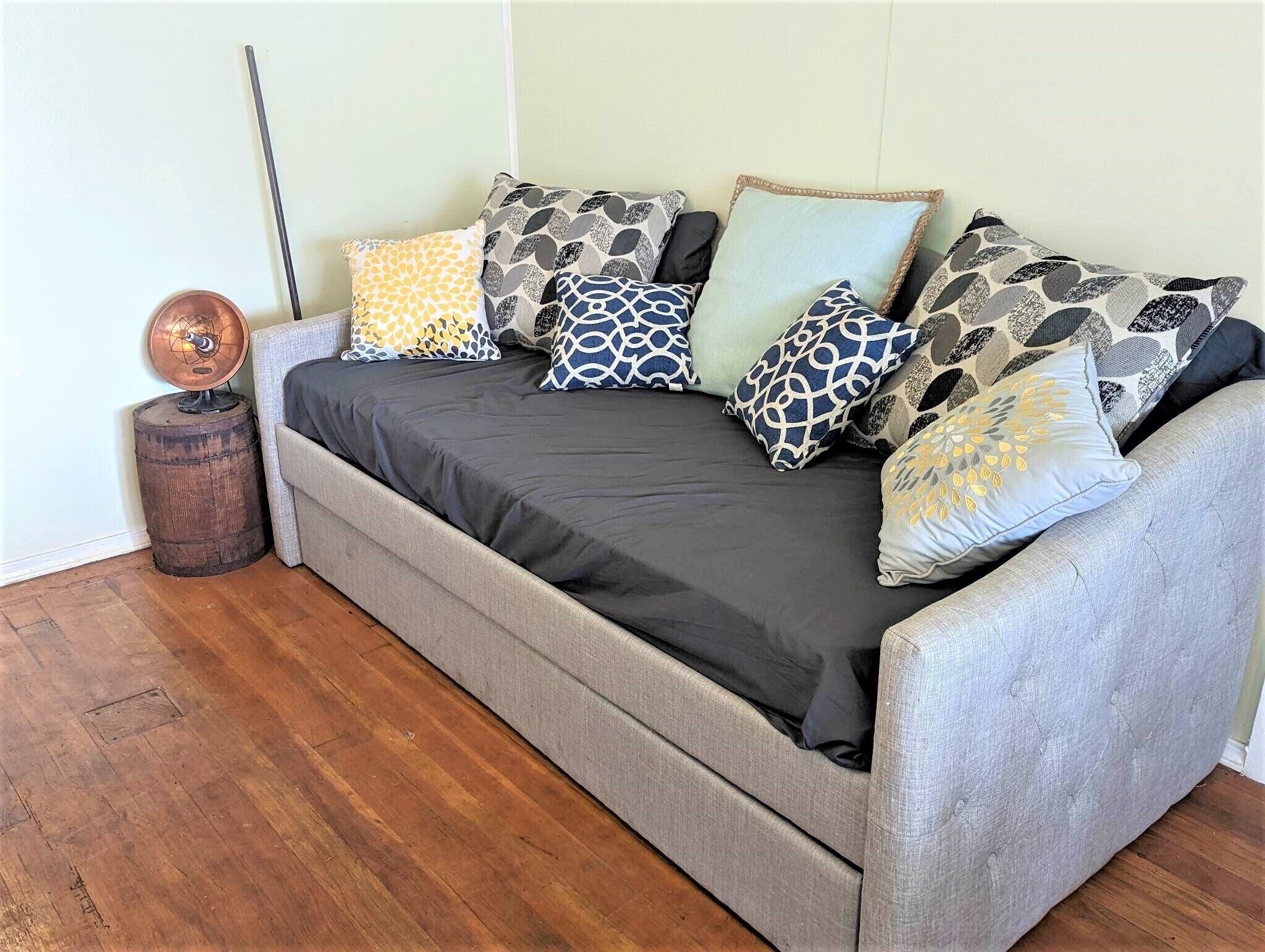 A grey sofa with a black cover and patterned cushions, a small wooden barrel, and a copper object on the floor.