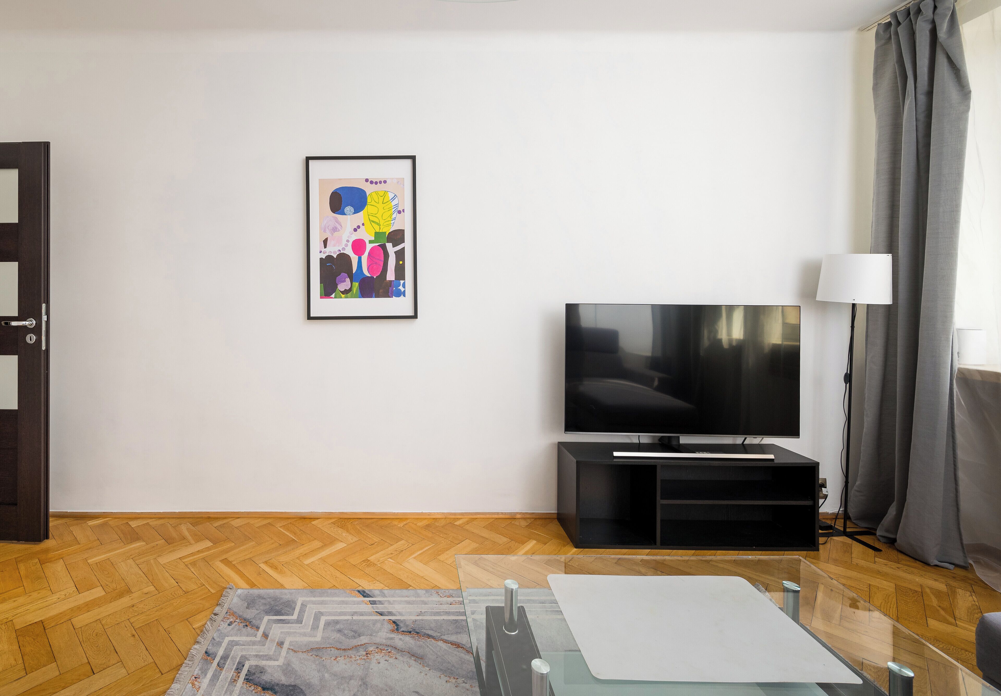 A modern living room with a flat-screen TV, a framed abstract painting, a glass coffee table, and a wooden herringbone floor.